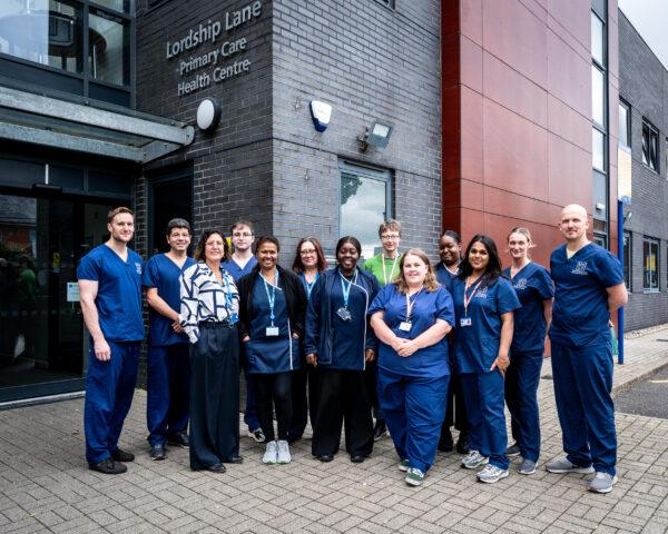 Team of healthcare staff, some wearing dark blue scrubs, some wearing civilian clothes standing outside GP practice, smiling.