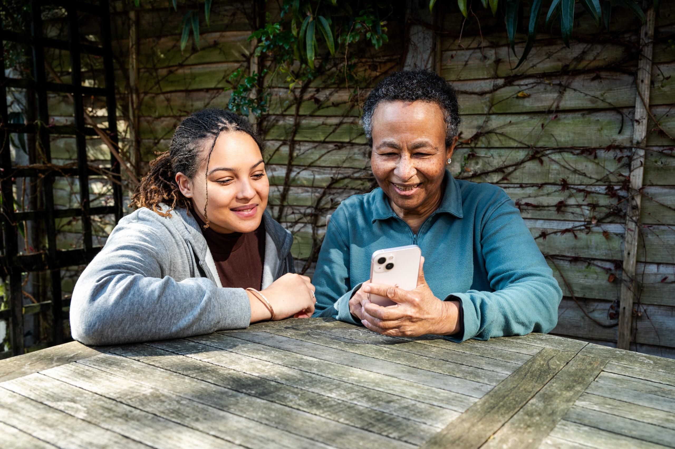 A young woman in a grey hoodie is sitting outside with an older lady, they are looking on a mobile phone at the NHS app 