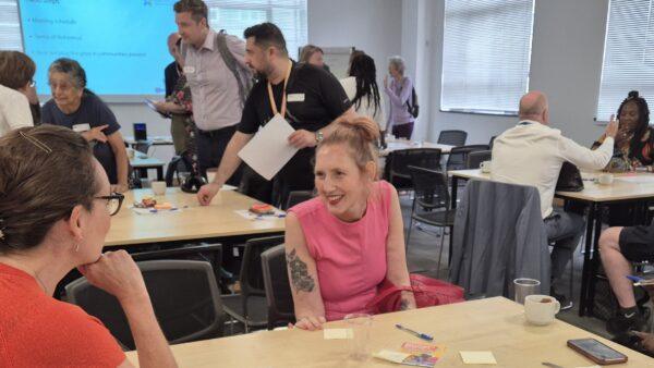 Two women talking and laughing while sat at a table, with a busy room of people talking in the background.